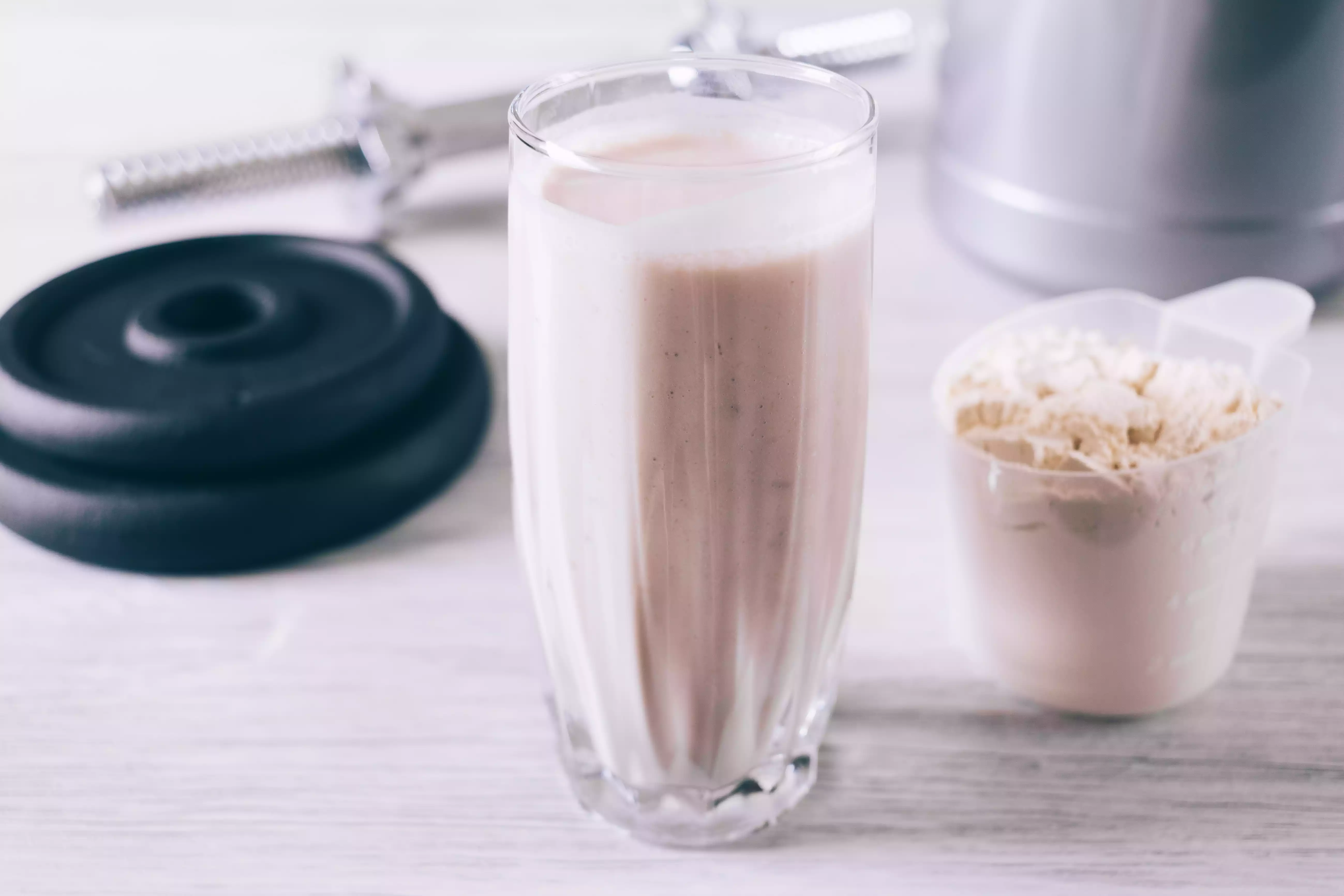 Scoop with protein powder, glass and dumbbell on a white wooden table